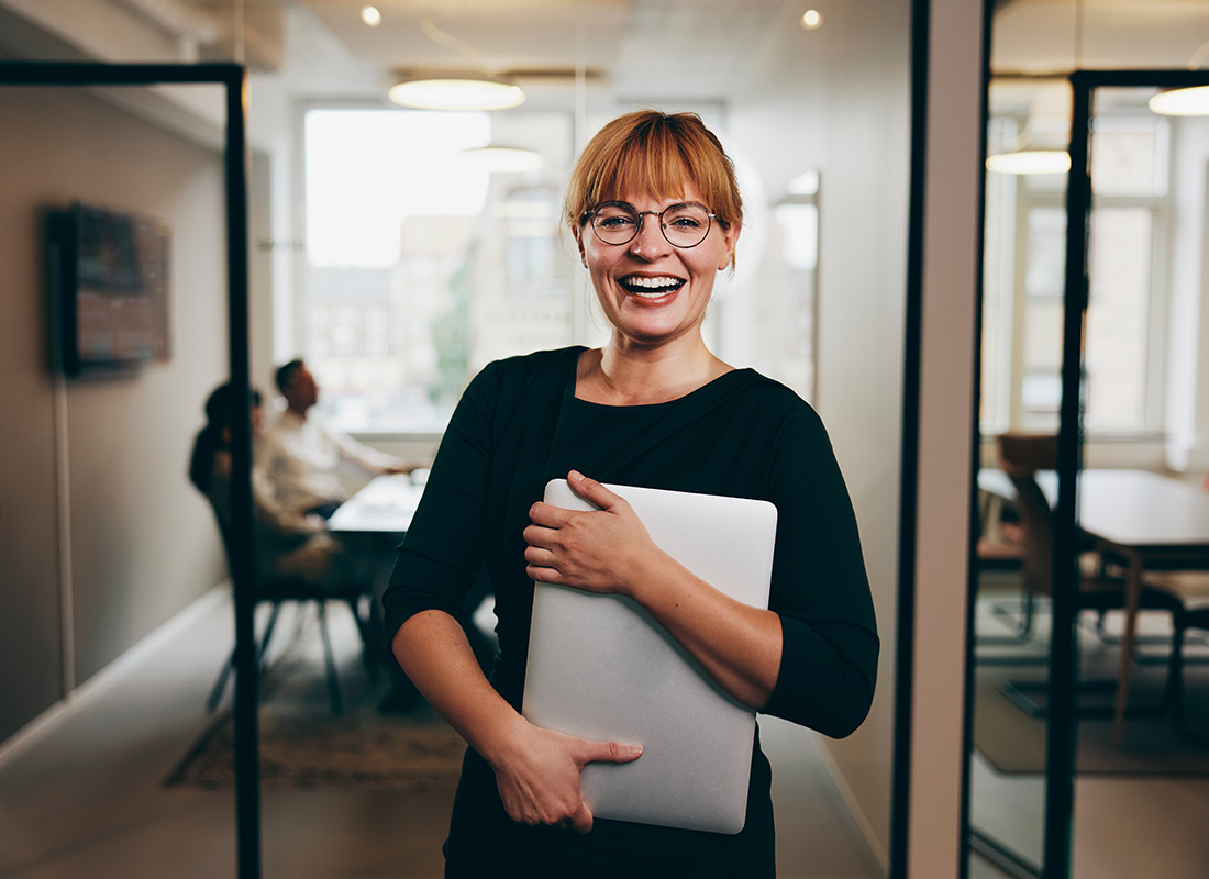 Business Insurance - Portrait of a Cheerful Middle Aged Business Woman Holding a Laptop While Standing in Front of a Conference Room with Other Employees Sitting in the Background