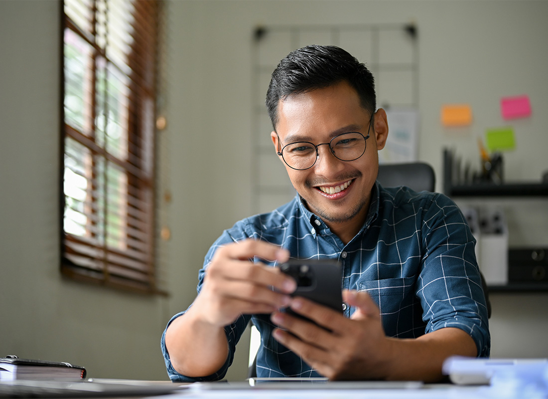Service Center - Closeup View of a Young Smiling Asian Businessman Wearing a Plaid Shirt Using a Phone While Sitting at his Desk
